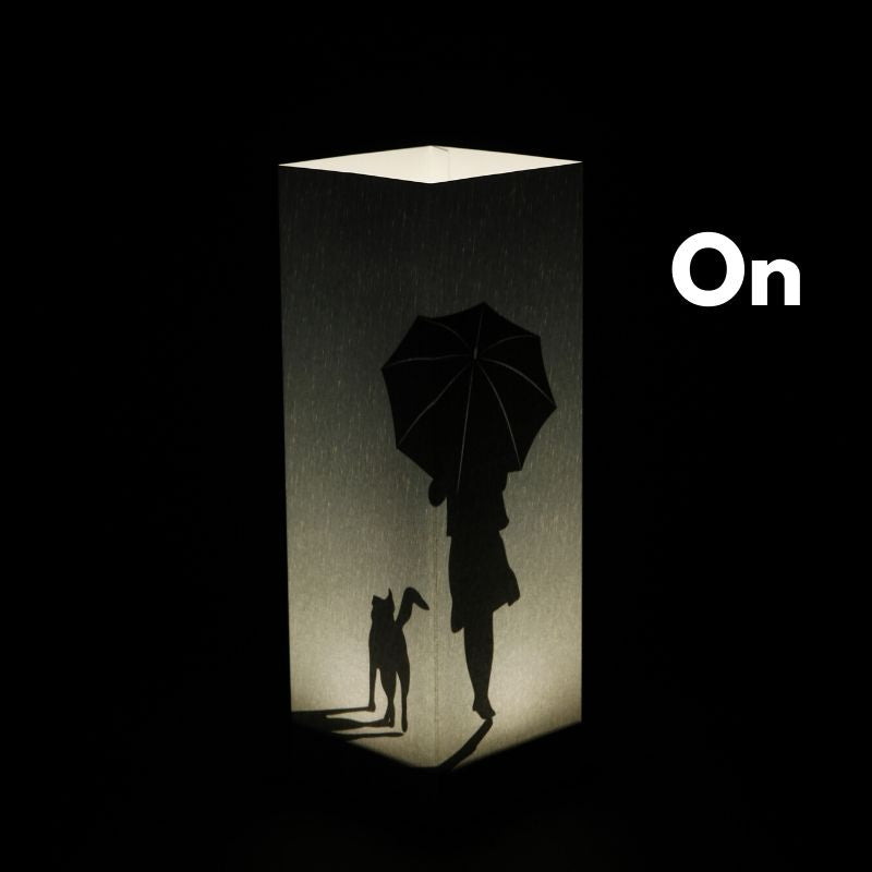 Zapnutá dekoračná tieňová LED lampa Rain Walker od LUXA London s postavou ženy kráčajúcou s psom v daždi